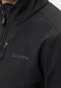 Veste softshell noire avec col montant, dotée d'une fermeture éclair frontale, logo sur la poitrine, et texture lisse et mate.