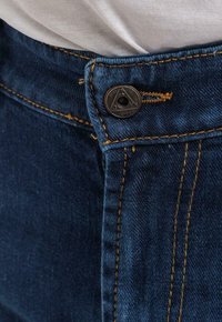 Džínové kalhoty s tmavě modrým odstínem, obsahující kontrastní žluté švy, kovové zapínání na knoflík a strukturovanou tkaninu.