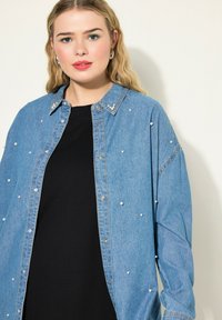 Camicia in denim azzurro chiaro con decorazioni in perle, chiusura a bottoni sul davanti e vestibilità rilassata. Sotto, una maglietta nera con scollo rotondo.