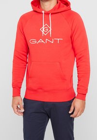 Sweat à capuche rouge en tissu doux avec une poche kangourou, arborant un logo blanc centré sur la poitrine et des cordons réglables sur la capuche.