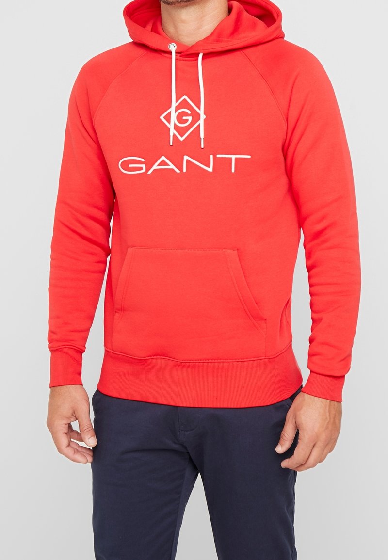 Sweat à capuche rouge en tissu doux avec une poche kangourou, arborant un logo blanc centré sur la poitrine et des cordons réglables sur la capuche.