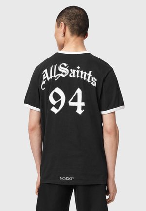 Mand med kort hår iført en sort T-shirt med hvid kant, der viser teksten "AllSaints 94" og "MCMXCIV" på ryggen.