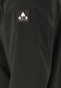 Schwarze Stoffjacke mit glatter Textur, die ein rechteckiges Logo-Patch mit der Aufschrift "WHISTLER" und einem stilisierten Bergdesign zeigt.