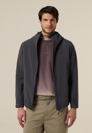 Mann trägt eine dunkelgraue Kapuzenjacke, ein violett verlaufendes T-Shirt, beige Hose und einen gemusterten Schal um den Hals gebunden, steht vor hellem Hintergrund.