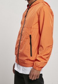 Orange lättvindjacka med huva, dragkedja framtill och svarta dragkedjor. Har elastiska muddar och sidofickor.