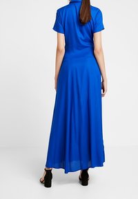 Sininen, lyhythihainen, maksimekko, joka on valmistettu sileästä kankaasta; siinä on kaulus ja hulmuava hame; yhdistetty mustiin korkokenkiin.