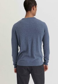 Blauer Strickpullover mit Rundhals-Ausschnitt, gerippten Ärmeln und Saum; glatte Textur; getragen mit dunkelgrauen Hosen.