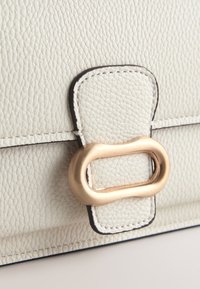 Gros plan d'un sac en cuir grainé blanc avec un rabat cousu et une boucle ovale dorée.