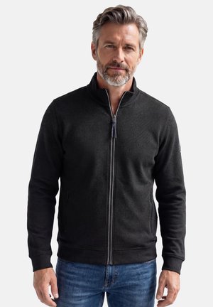 Mann mit grauem Haar und Bart, der eine schwarze Jacke mit Reißverschluss und blaue Jeans trägt, steht vor einem schlichten hellen Hintergrund.