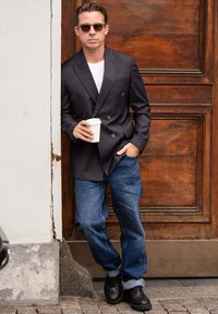 Sort dobbeltradet blazer over en hvid skjorte, parret med blå denim jeans og sorte sko, mens der holdes en kaffekop, mod en trædør.