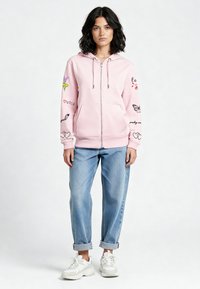 Svijetloruža majica s kapuljačom na patentni zatvarač, s kapuljačom, ukrašena šarenim grafikama i tekstom, u kombinaciji s rolanim trapericama i bijelim tenisicama.