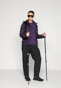 Giacca isolante viola sopra una maglietta viola, pantaloni da trekking neri e bastoncini da trekking. Occhiali da sole e zaino nero. Texture liscia e lucida.