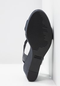 Semelle de sandale noire avec bande de roulement texturée et logo "Tommy Jeans", présentée en angle contre un mur et un sol blancs en coin.