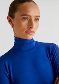 Blauer Rollkragenpullover aus glattem Stoff, mit tailliertem Schnitt und feiner Textur. Das Modell trägt einen kleinen silbernen Creolenring.