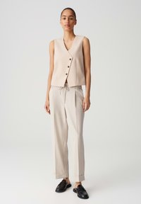 Beige mouwloos vest met knoopsluiting, gecombineerd met lichtbeige broek met trekkoord. Zachte textuur, relaxte pasvorm en plooien bij de broekspijpen.