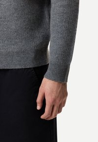 Rechte Hand und Handgelenk einer Person, die einen grauen, strukturierten Pullover und schwarze Hosen trägt, vor einem schlichten weißen Hintergrund.