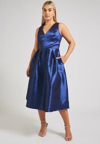 Sininen satiini mekko, jossa on V-pääntie, hihaton muotoilu ja rakenteelliset laskokset. Mekossa on sivutaskut ja valunut A-linjan muoto.