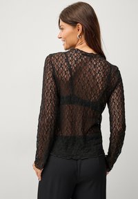 Sort blonde langærmet top med en gennemsigtig, indviklet blomsterprint, scalloped kant og tekstureret finish. Bagfra ses det tætsiddende design.