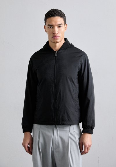 Casaco preto com fecho éclair e capuz, feito de material leve, com um padrão texturado subtil e punhos elásticos, combinado com calças cinza claro.