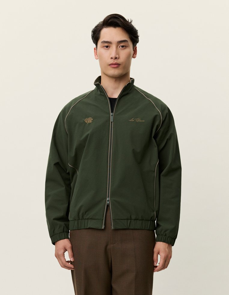 Tmavozelená bunda na zips s textúrovanou tkaninou, elastickými manžetami a zlatou vyšitou logom na hrudi. Zdobená svetlým prešívaním.