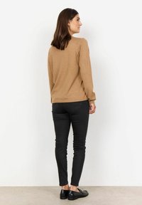 Maglione marrone chiaro con maniche lunghe, abbinato a jeans skinny neri. Il maglione ha un orlo arrotondato, mentre i jeans presentano una vestibilità classica. Scarpe nere completano l'outfit.