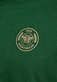 Tissu de sport vert avec logo d'abeille beige entouré de branches de laurier et du texte «1923 F.C. Sportswear» en cercle.