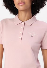 Vaaleanpunainen polo-paita, jossa on kaulus, neljä nappia ja brodeerattu "New York Tommy Hilfiger" -logo. Kankaan pinta on teksturoitu.