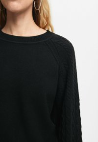 Maglione nero a maglia con collo rotondo, caratterizzato da un design a trecce alle maniche e una texture liscia. Nessun motivo o accento visibile.