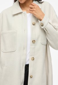 Ανοιχτό καφέ μάλλινο παλτό με γιακά, δύο μπροστινές τσέπες και μεγάλους κόμπους. Μαλακή υφή και χαλαρή εφαρμογή, σχεδιασμένο για άνεση.