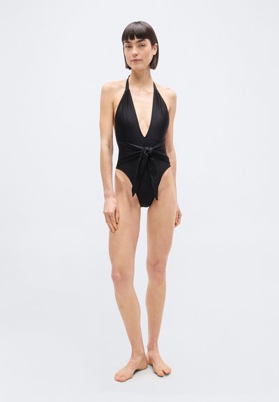 Γυναίκα που στέκεται ξυπόλητη φορόντας μαύρο ολόσωμο μαγιό με βαθύ V και δέσιμο μπροστά, σε απλό ανοιχτό φόντο.