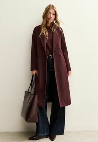 Cappotto lungo bordeaux con rever a punta, abbinato a un maglione coordinato, jeans a gamba larga scuri e una borsa di pelle testurizzata. Scarpe con punta a cono.