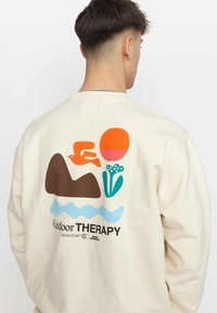 Crème sweatshirt met een kleurrieke grafiek op de achterkant, afgebeeld met een vogel, berg, zon, bloemen en golven, met zwarte tekst eronder.