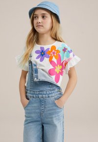 Witte T-shirt met kleurrijke bloemenprint; blauwe denim overalls met verstelbare bandjes; blauwe bucket hat; details met ruches bij de mouwen.