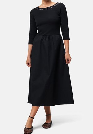 Vrouw die een zwarte midi-jurk draagt met driekwart mouwen, een met studs versierde halslijn en zwarte mesh enkellaarzen tegen een effen achtergrond.