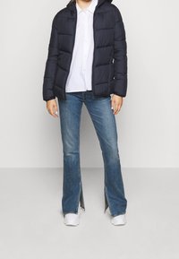 Veste rembourrée bleu marine avec capuche, fermeture éclair à l'avant et texture matelassée. Associée à un jean léger évasé bleu et des baskets blanches, créant un look décontracté.