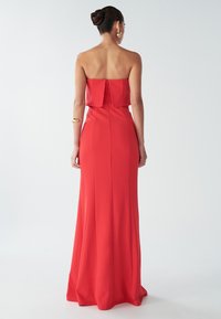 Punainen, olkaimeton iltapuku, jossa on istuva yläosa ja virtaava hame. Sujuva kangas, pitkä siluetti ja piilotettu takavetoketju.