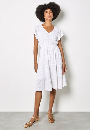 Mujer con cabello rizado vestida con un vestido blanco de manga corta y escote en V, con detalles de ojales y sandalias de tacón doradas con tiras, de pie.