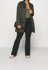 Olivgrön mockajacka i kombination med en grå topp, svarta byxor och vita sandaler. Håller en svart kuvertväska. En enkel och stilren design.