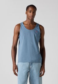 Débardeur en coton bleu avec des emmanchures larges et un col rond, arborant un petit logo rouge Levi's sur le côté gauche. Porté avec un jean bleu clair.