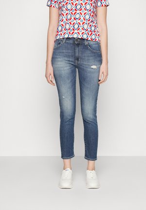 Kvinde iført falmede blå slim-fit jeans med små slidte pletter, kombineret med hvide sneakers og en mønstret kortærmet top.