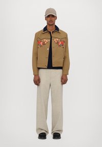 Khaki bunda s květinovou výšivkou, tmavě modrý límec, knoflíky, kombinovaná s světle béžovými širokými kalhotami a černými botami.