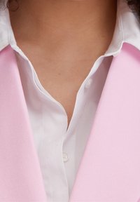 Gros plan sur une personne portant une chemise blanche boutonnée sous un blazer rose clair, montrant le cou et la partie supérieure de la poitrine.