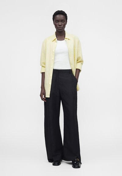 Γυναίκα με κοντά μαλλιά που φοράει ένα ανοιχτό κίτρινο πουκάμισο με κουμπιά πάνω από λευκό τοπ, μαύρο φαρδύ παντελόνι και μαύρα παπούτσια, στέκεται με το ένα χέρι στην τσέπη.