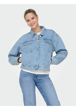 Lachende vrouw met blond haar in een lichtblauwe spijkerjas en bijpassende jeans, staand met de handen in de zakken tegen een witte achtergrond.