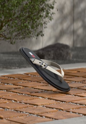 Sandale noire à bride avec boucle beige flottant au-dessus d'un sol en carreaux de bois et galets dans un cadre extérieur avec des arbustes verts et un mur en pierre en arrière-plan.