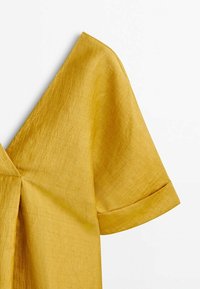 Chemise en lin jaune à manches courtes avec un revers de manche relevé et des détails de coutures visibles sur fond blanc.