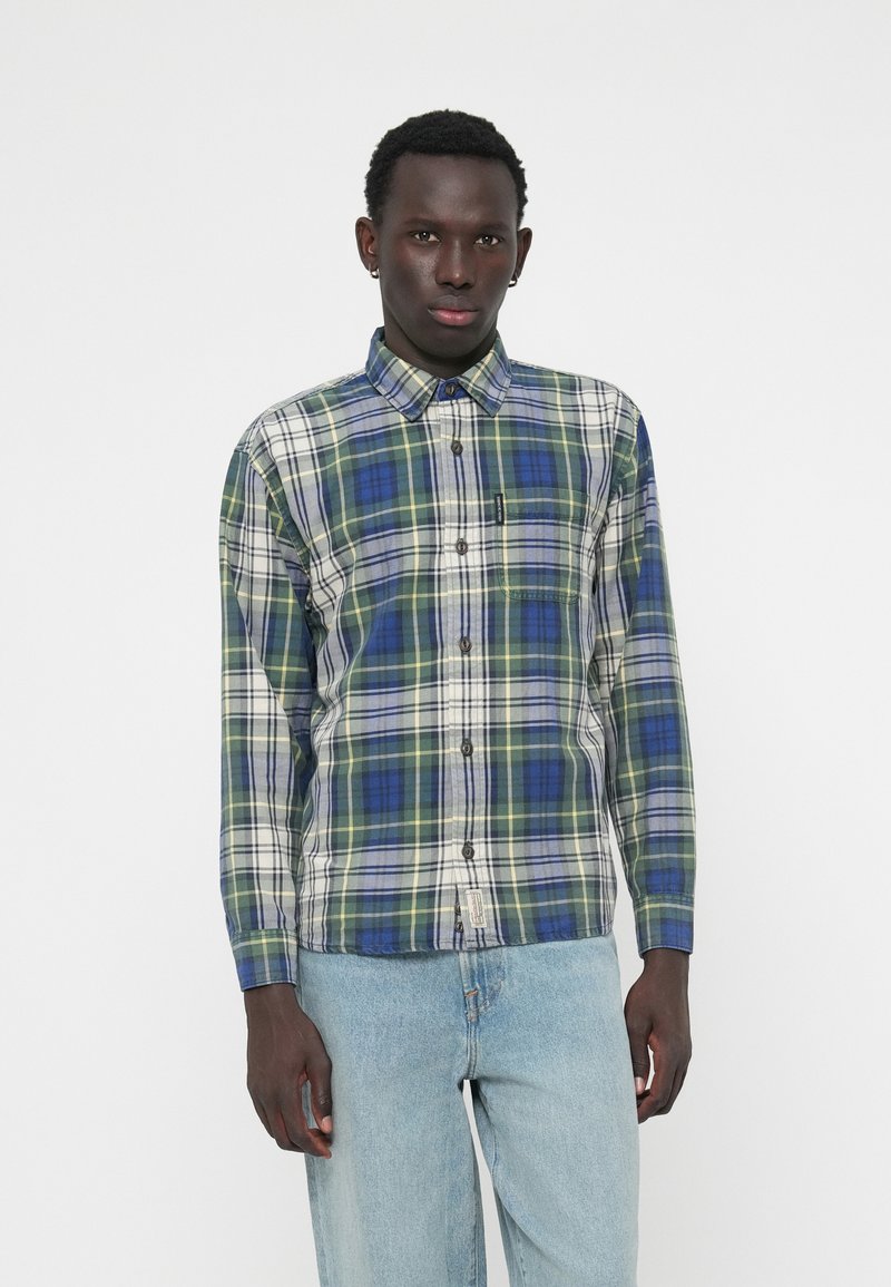 Chemise à manches longues à carreaux bleu, vert et crème. Elle possède un col, des boutons sur le devant et une poche poitrine. Portée avec un jean bleu clair.