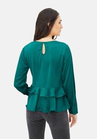 Blusa de satén en color verde azulado con mangas largas, dobladillo con volantes y puños abotonados. Presenta un pequeño detalle en forma de ojal en el escote trasero.