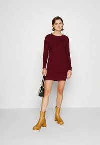 Γυναίκα που στέκεται φορώντας ένα βουργουνδί πλεκτό φόρεμα με μακριά μανίκια, καφέ μποτάκια μέχρι τον αστράγαλο, κρατώντας μια μαύρη τσάντα χειρός με χρυσές αλυσίδες.