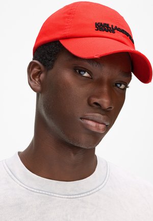 Hombre joven negro que lleva una gorra roja con el logo "KARL LAGERFELD JEANS" y una camiseta blanca de cuello redondo, mirando ligeramente hacia la derecha.